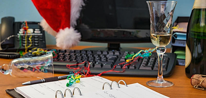 Schreibtisch mit aufgeschlagenem Kalender, Monitor und Tastatur, Luftschlangen, Sekt und Weihnachtsmütze.