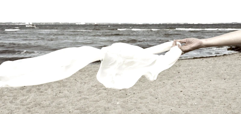 Eine Hand hält ein weißes Tuch, das am Meeresstrand im Wind weht.